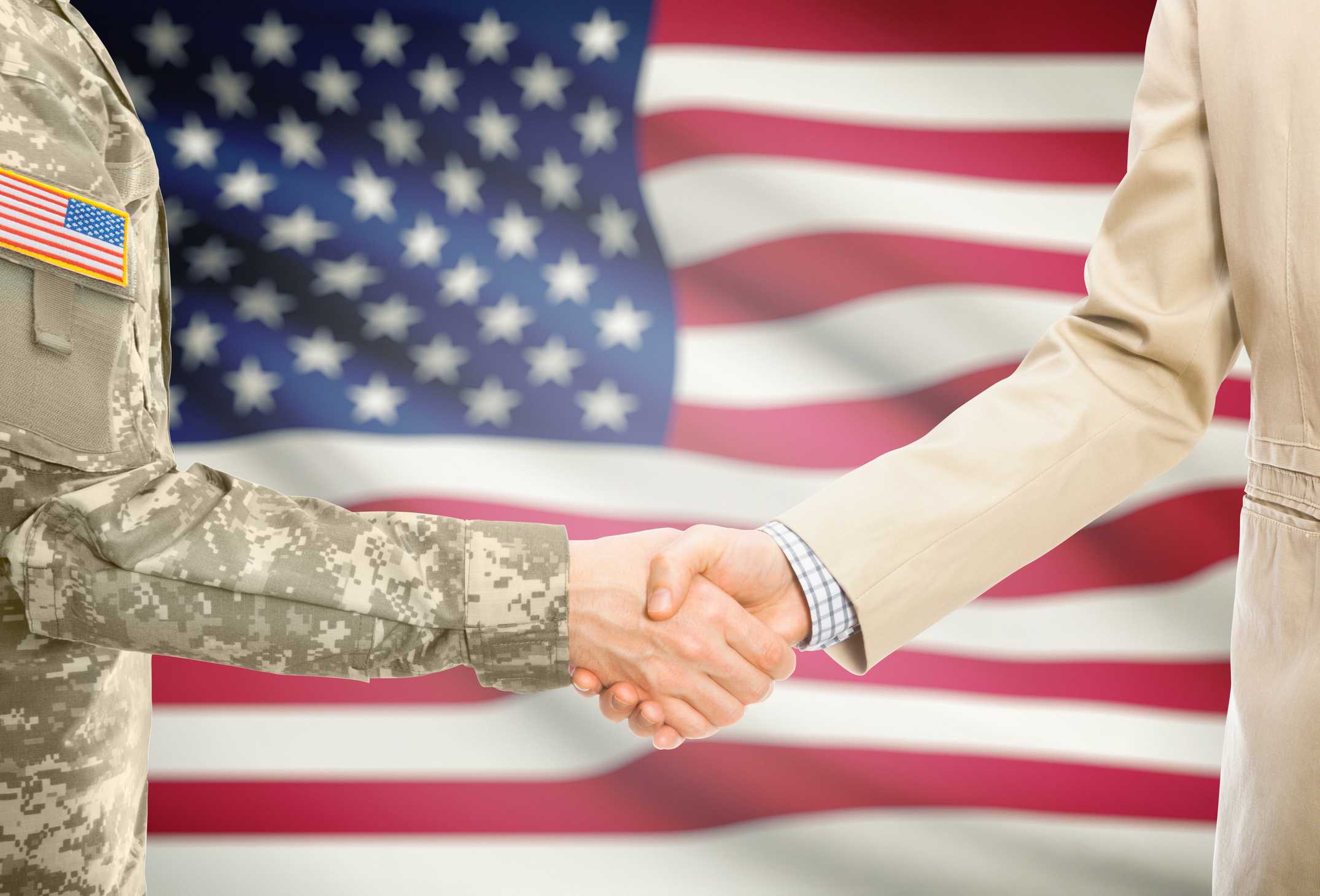American soldier in uniform and civil man in suit shaking hands in front of a flag.